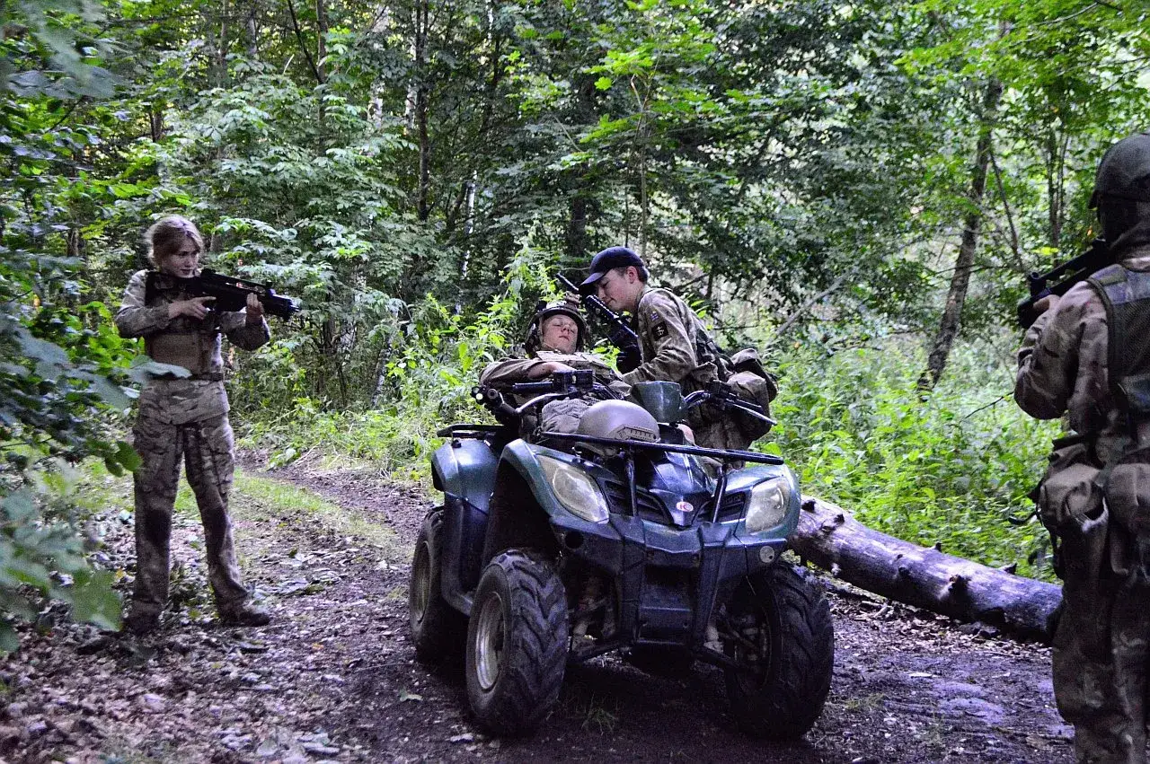 Trzech graczy ASG w kamuflażu, jeden na quadzie, dwóch z replikami broni, w lesie.