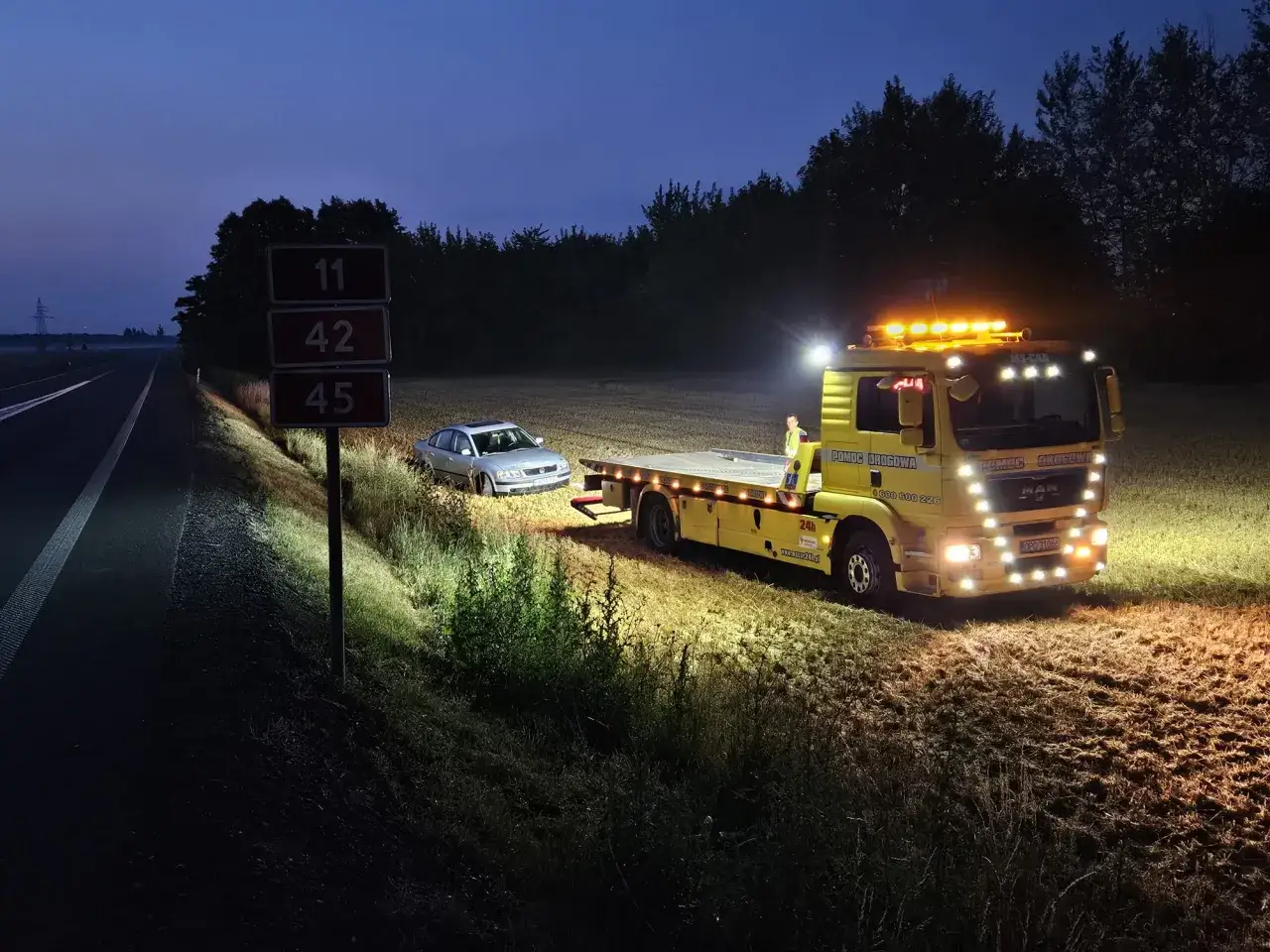 Nocna pomoc drogowa dla srebrnego samochodu na poboczu. Żółty holownik z włączonymi światłami zapewnia wsparcie.