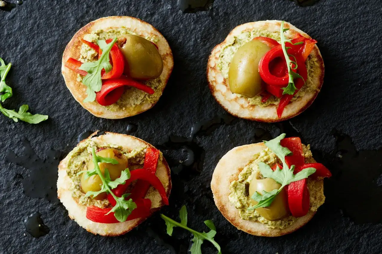 Vier kleine, runde Snacks mit Pesto, gr&uuml;ner Olive, roten Paprikastreifen und Rucola. K&ouml;stliches fingerfood vegetarisch.