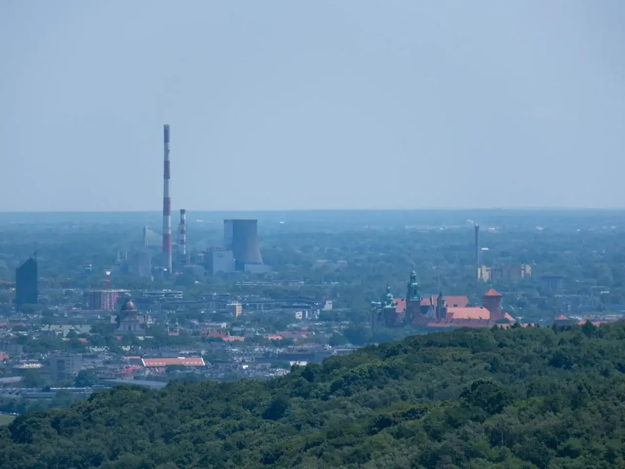Rozległa panorama Krakowa z dominującymi wieżami Wawelu i kominami elektrociepłowni na horyzoncie.
