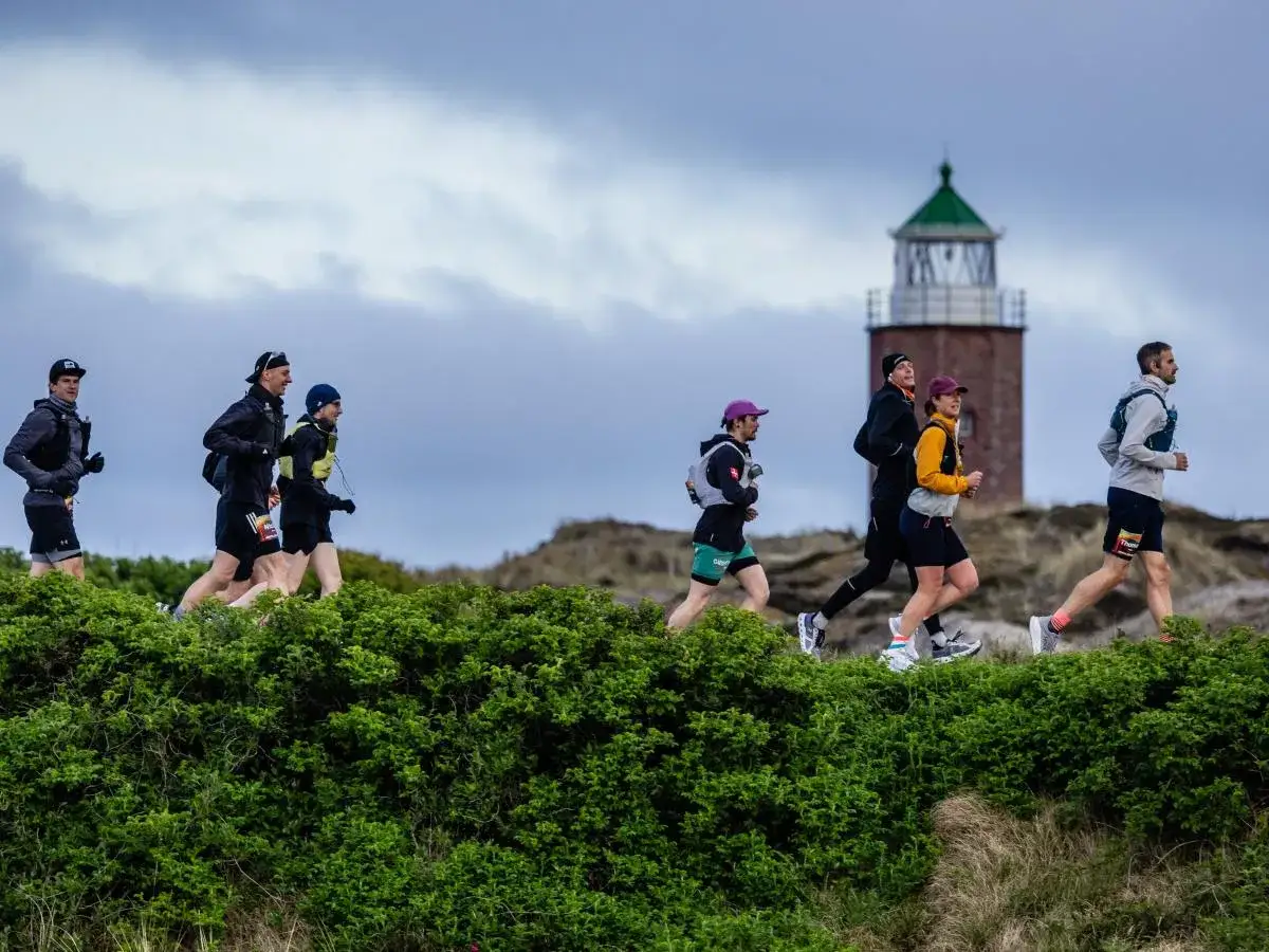 Ultramarathonläufer laufen an einem Leuchtturm vorbei durch eine grüne, hügelige Landschaft.