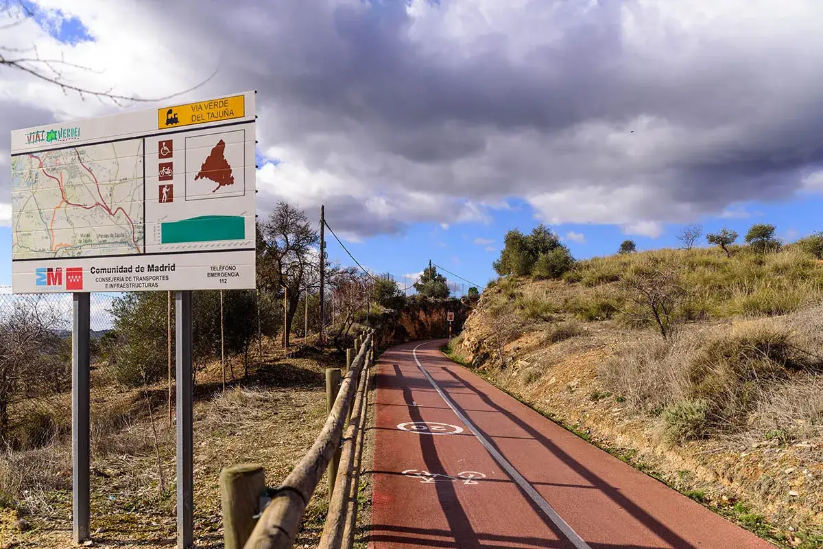 Cartel informativo de la v&iacute;a verde del Taju&ntilde;a. Un camino rojo para bicicletas y peatones serpentea por un paisaje seco y soleado.