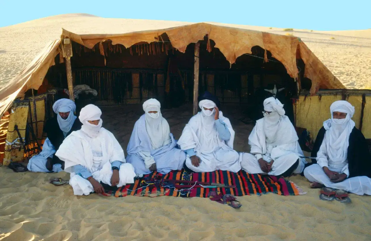 Sieben M&auml;nner eines Berberstamms in der Sahara sitzen vor einem Zelt auf einer bunten Decke.