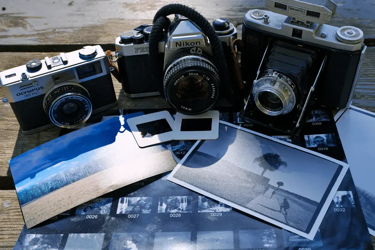 Kolekcja starych aparat&oacute;w i odbitek. Widać Olympus 35 RC, Nikona FE2, aparat składany Zeiss Ikon i slajdy. To piękne zdjecia analogiem.