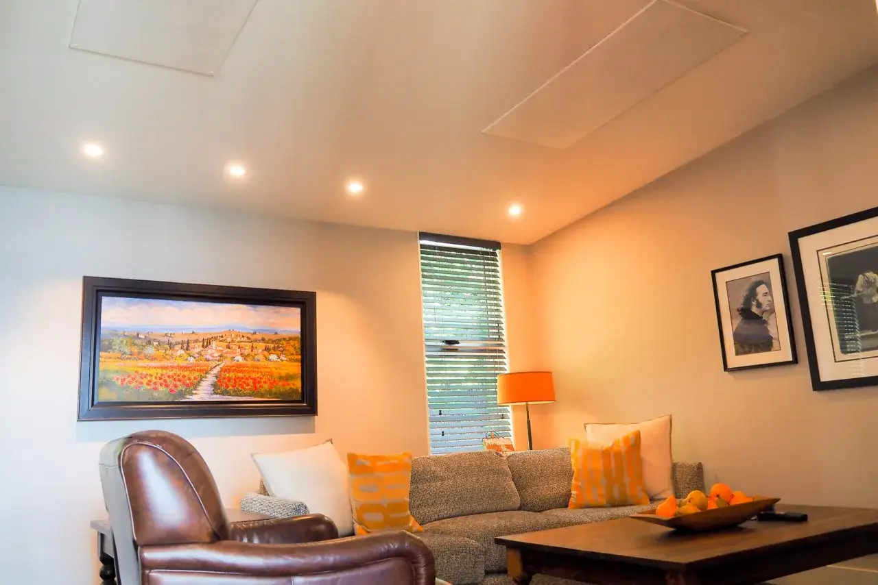 Salon confortable avec un tableau de paysage, un fauteuil en cuir et un canap&eacute;. Un panneau discret au plafond sugg&egrave;re un syst&egrave;me de chauffage infrarouge.