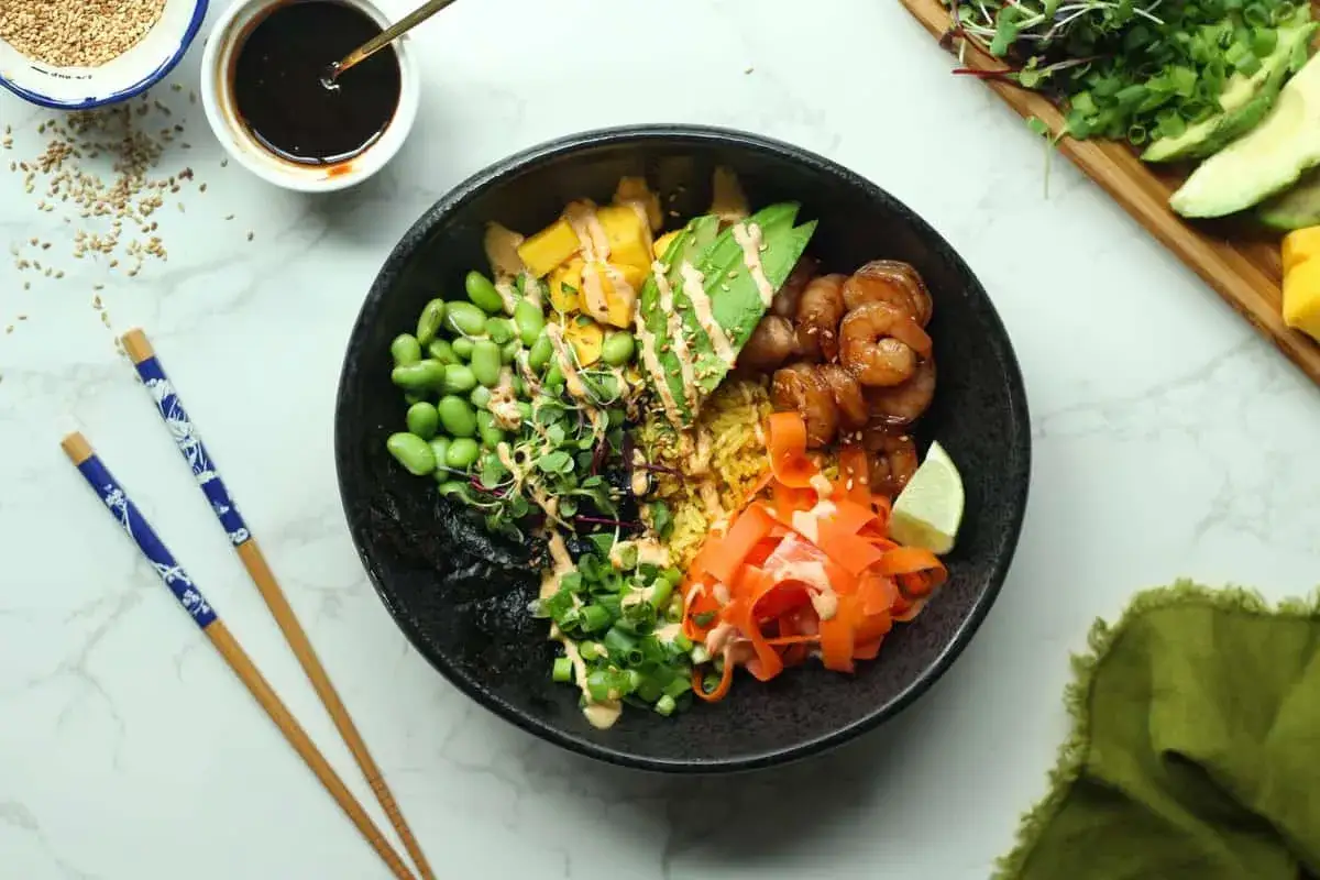 Kolorowy poke bowl z krewetkami, mango, awokado, edamame i startą marchewką, polany sosem.