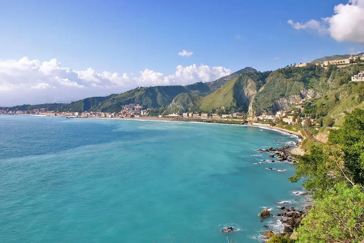 La costa di Giardini Naxos si estende sotto colline verdi, con acque turchesi che lambiscono rocce e una spiaggia sabbiosa.