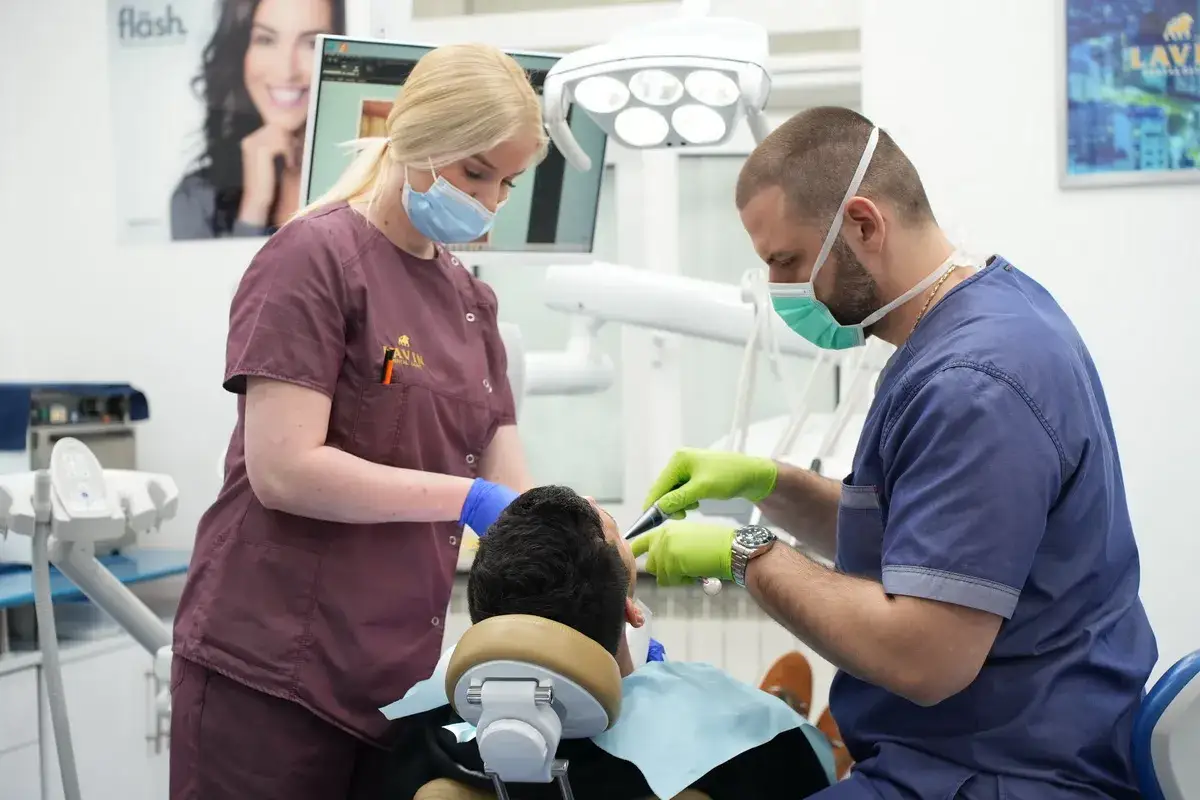 Dentysta i asystentka w maseczkach i rękawiczkach wykonują zabieg. Pacjent leży na fotelu, czując się komfortowo dzięki sedacji.