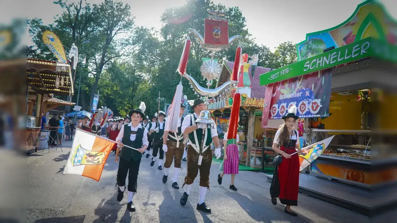 Kinder und Erwachsene in Tracht marschieren beim Volksfest Freising. Fahnen und ein festlicher Wagen ziehen vorbei.