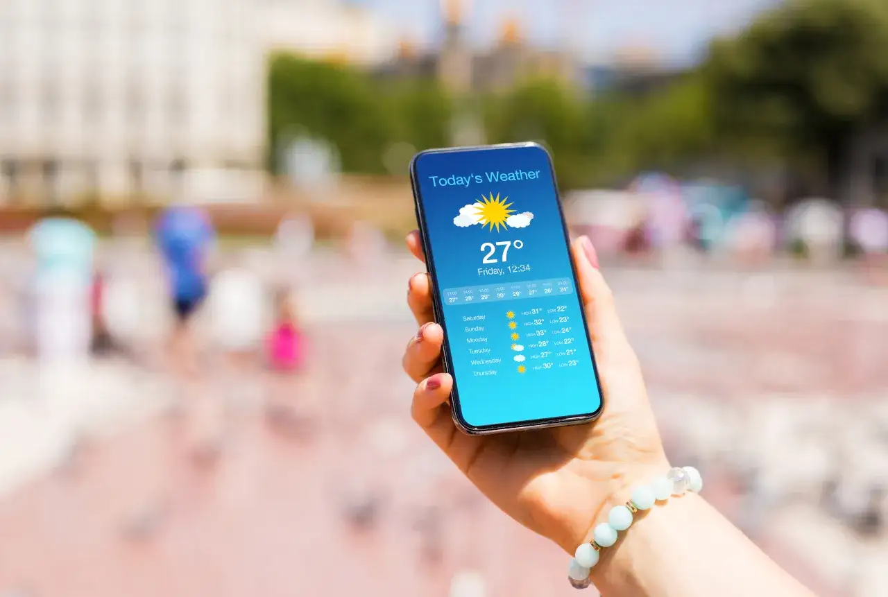 Dłoń trzyma smartfon z wyświetloną prognozą pogody, pokazującą 27°C i zapowiedź słonecznego dnia. To jedna z najlepszych aplikacji pogodowych, która pomaga planować aktywności.