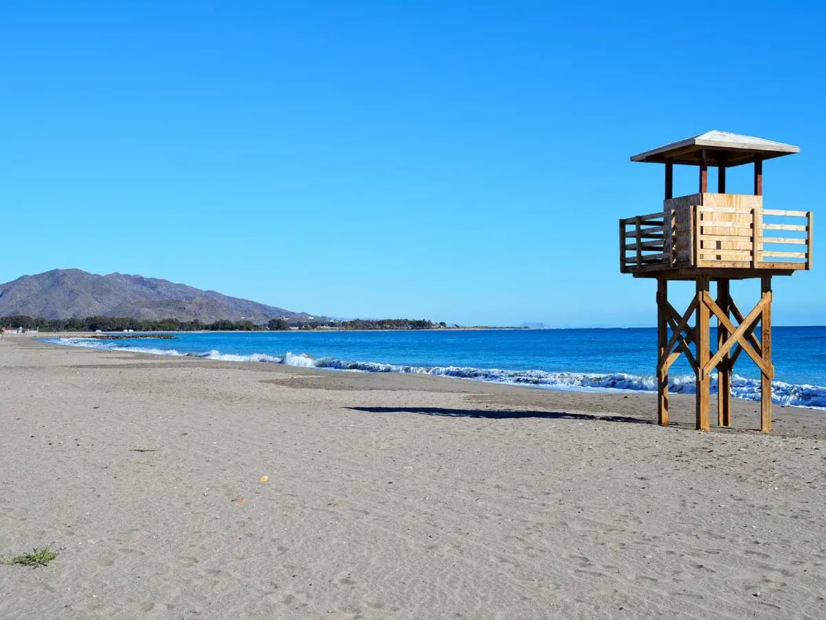 Torre de vigilancia de madera en la vera playa, con el mar azul y montañas al fondo.