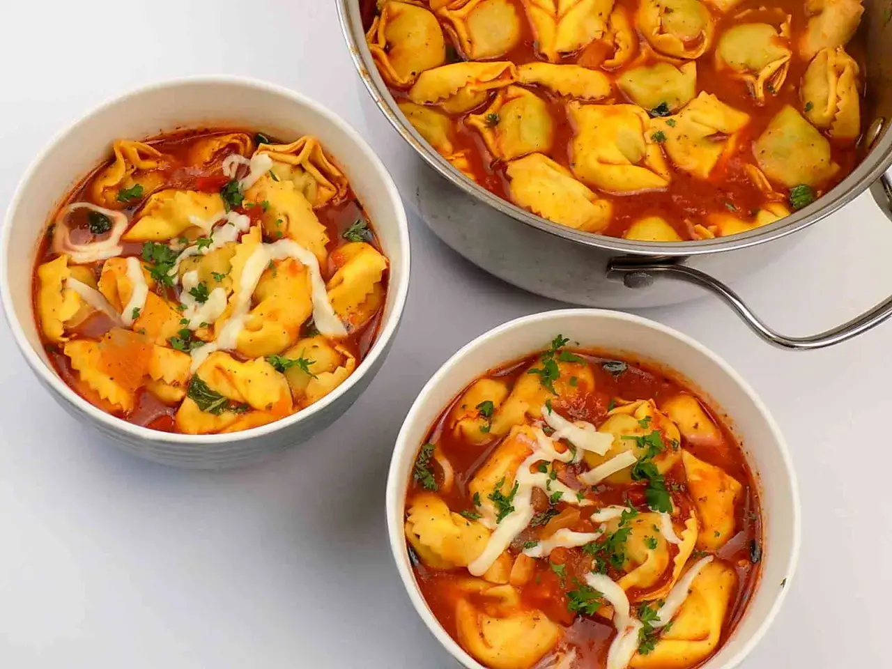 Dwie miseczki i garnek pełne aromatycznej zupy z tortellini w sosie pomidorowym, posypanej serem i natką pietruszki.