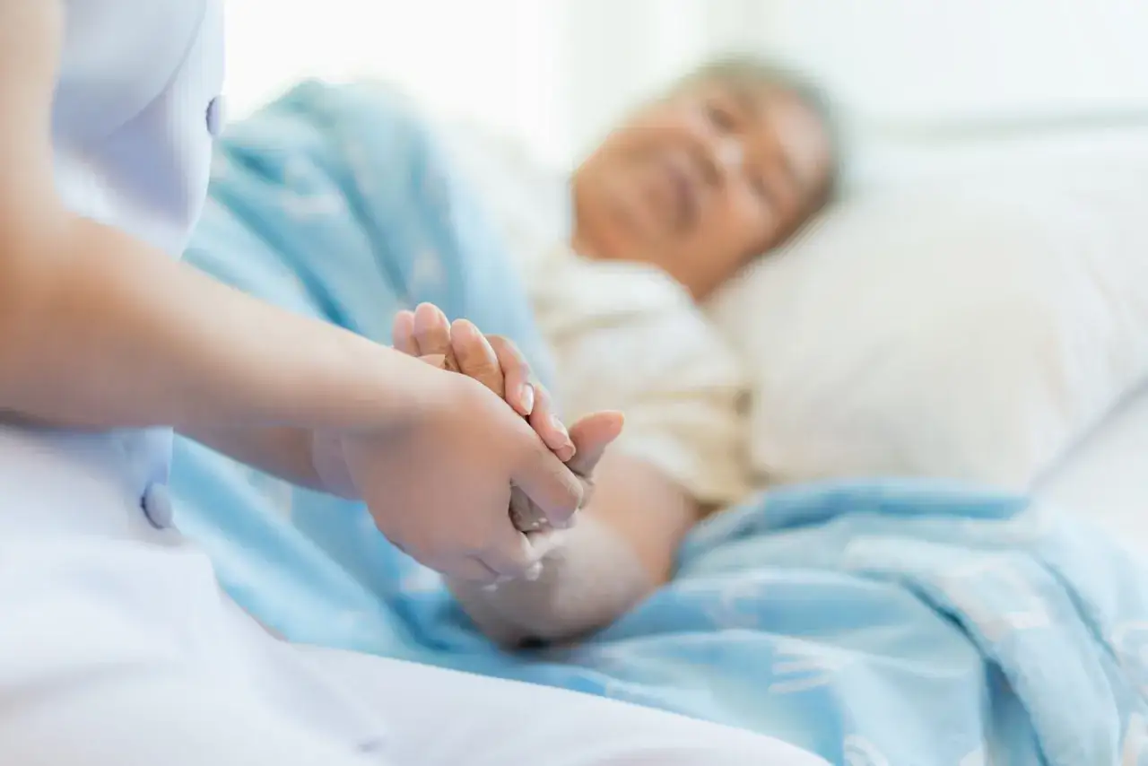 Pielęgniarka trzyma za rękę pacjentkę, kt&oacute;ra leży w ł&oacute;żku. Widać delikatne odleżyny na plecach pacjentki, kt&oacute;re wymagają troskliwej opieki.