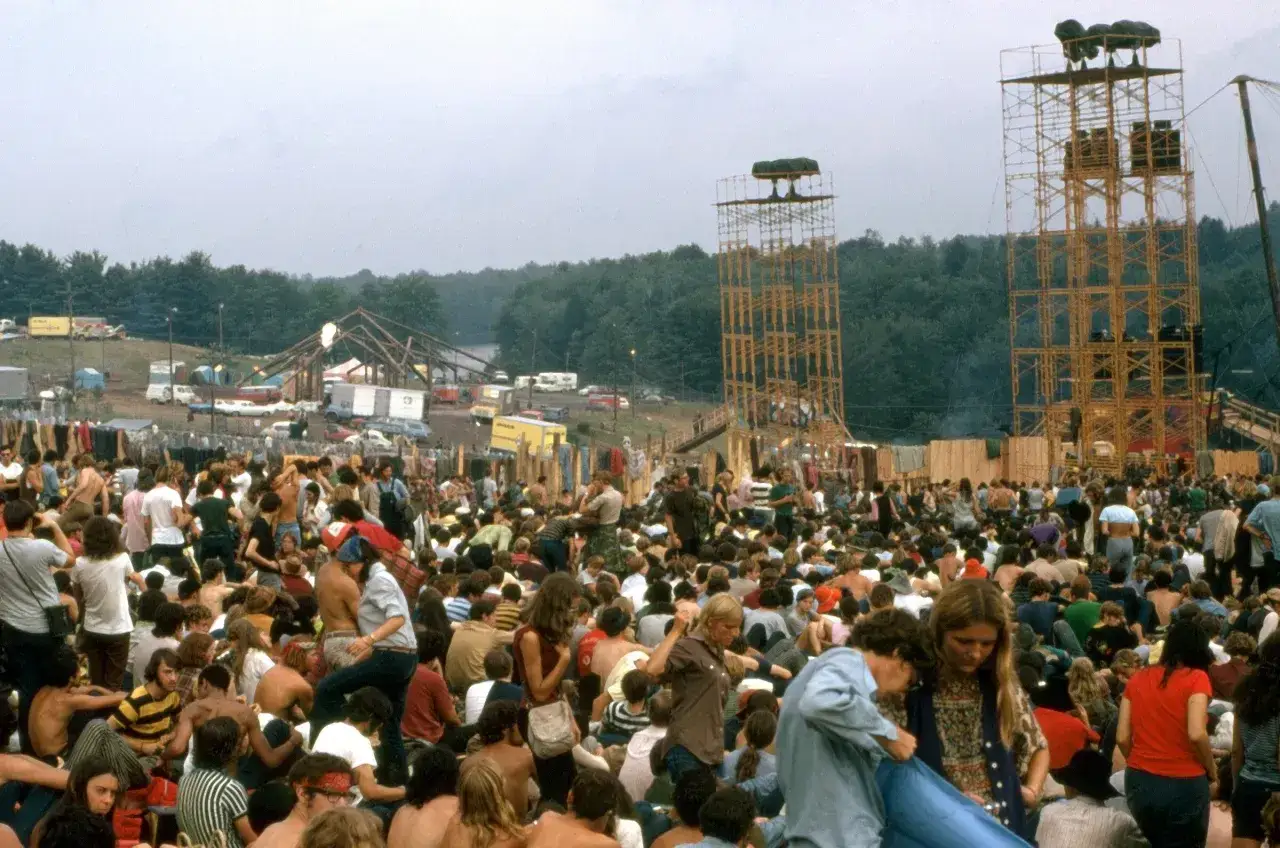 Tłum ludzi na festiwalu muzycznym, gdzie jest Woodstock. W tle widać konstrukcje sceniczne i las.