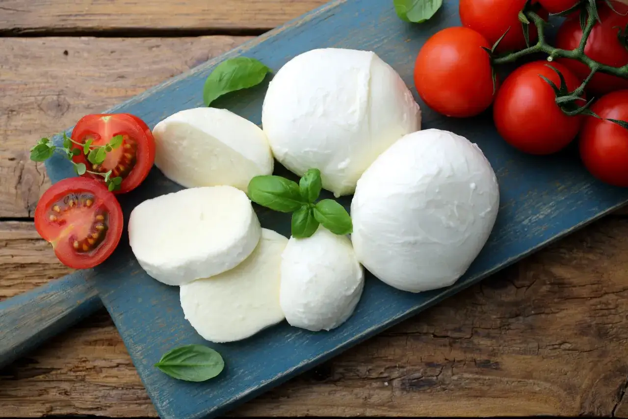 Bocconcini di mozzarella fresca, pomodorini maturi e foglie di basilico su un tagliere blu.