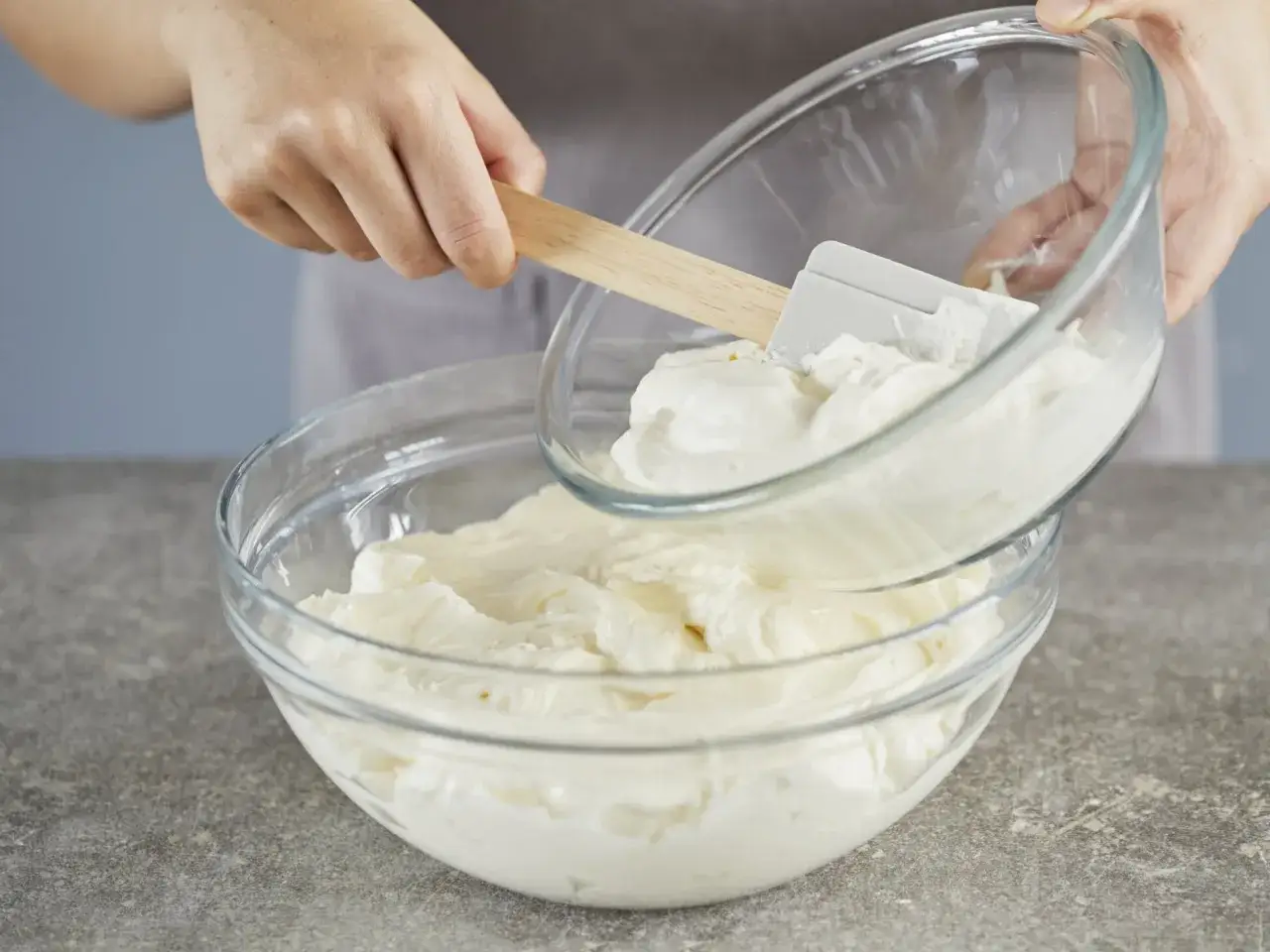 H&auml;nde geben mit einem Spatel eine Sch&uuml;ssel mit geschlagener schwerer Sahne in eine gr&ouml;&szlig;ere Sch&uuml;ssel mit cremiger Masse.