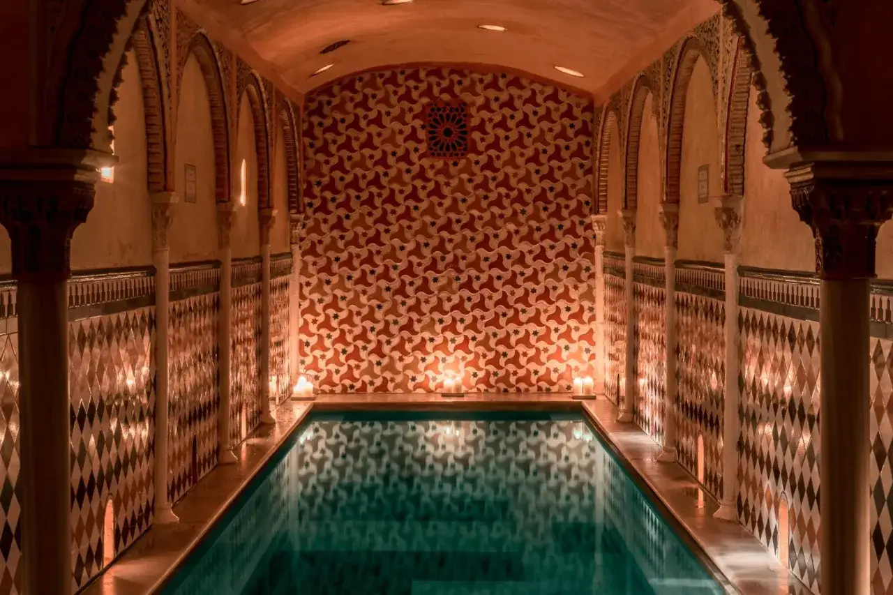 Ein luxuri&ouml;ser Hammam in Granada mit einem Pool, umgeben von kunstvollen Kacheln und S&auml;ulen, beleuchtet von Kerzen.