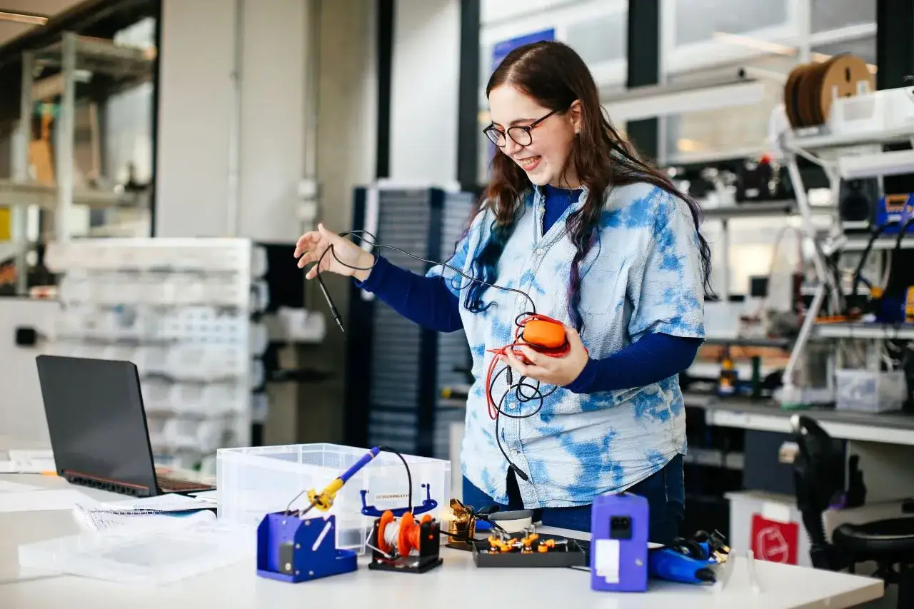 Junge Frau mit Brille lacht und h&auml;lt ein orangefarbenes Ger&auml;t mit Kabeln. Sie arbeitet in einem Labor, ein Beispiel f&uuml;r kreative Berufe.