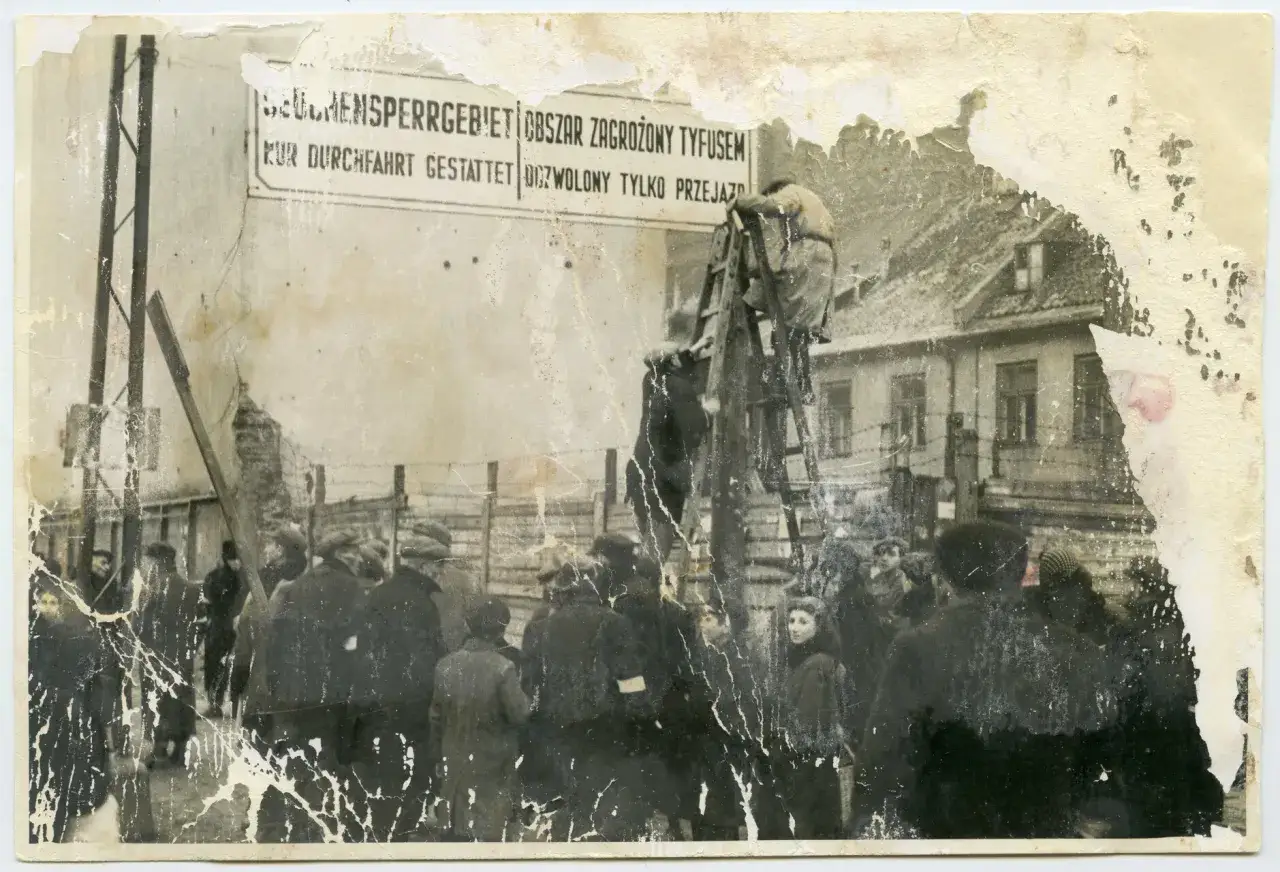 Ostrzeżenie o obszarze zagrożonym tyfusem w 1941 roku. Ludzie stoją za drutem kolczastym, w tle budynki.
