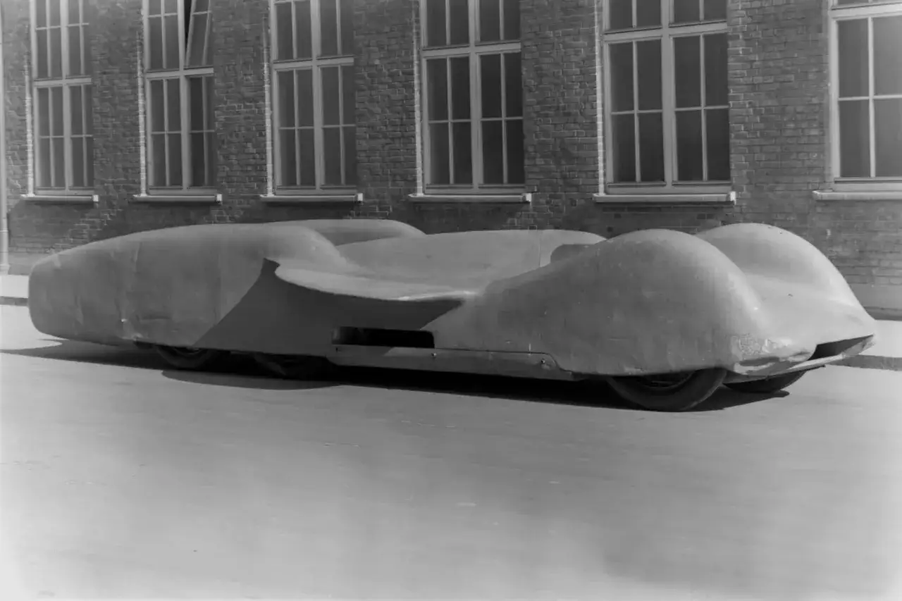 Der aerodynamische Mercedes-Benz T 80, ein Rekordfahrzeug, steht vor einer Backsteinmauer mit gro&szlig;en Fenstern.