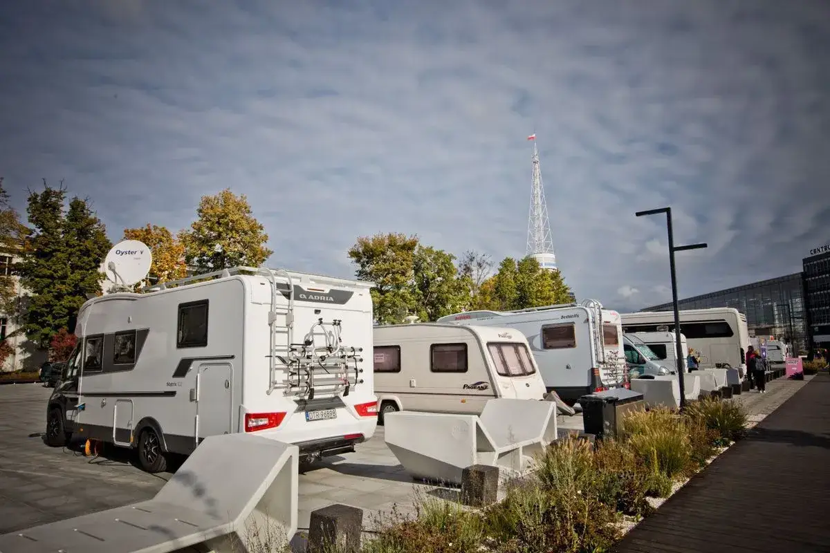 Zlot caravaningowy w mieście. Rzędy kamperów i przyczep kempingowych zaparkowane obok siebie, z wieżą radiową w tle.