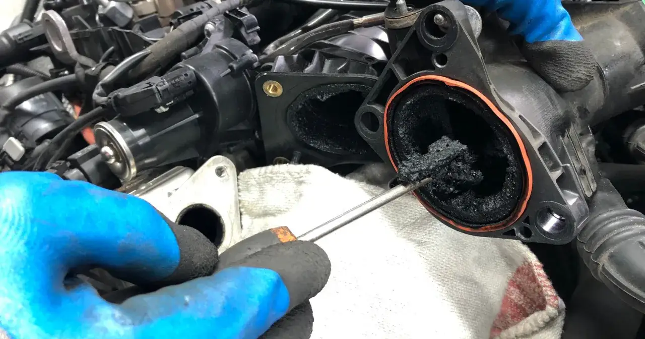 Gęsty nagar blokuje dolot silnika. Mechanik w niebieskich rękawiczkach próbuje go usunąć.