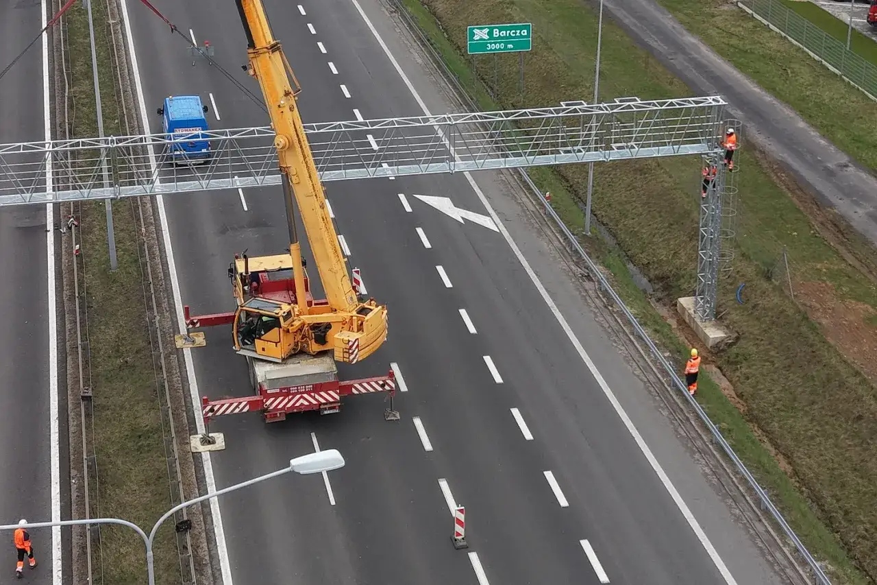 Montaż bramownicy do odcinkowego pomiaru prędkości S7. Dźwig pomaga pracownikom w instalacji konstrukcji nad autostradą.