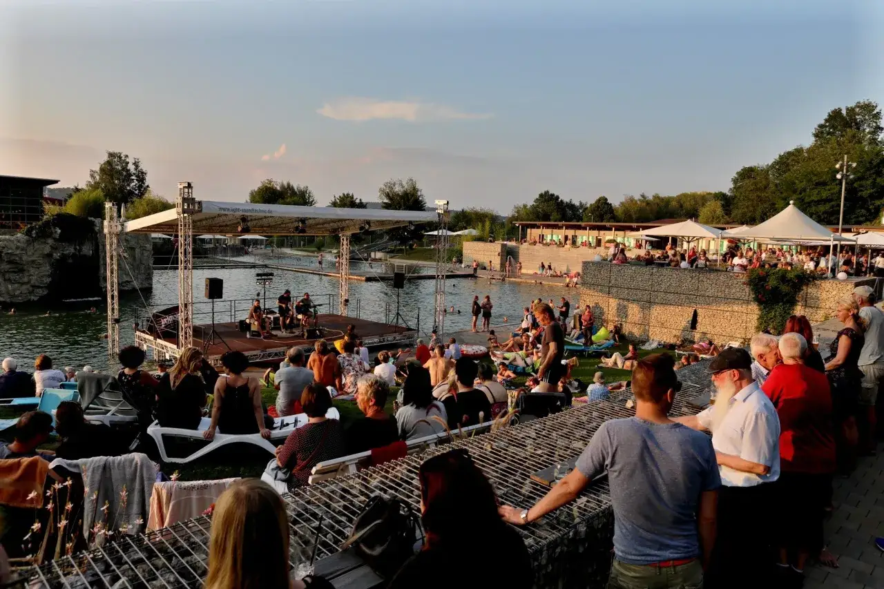 Sommerabend am Badesee Schorndorf: Menschen entspannen, baden und genie&szlig;en ein Konzert am Wasser.