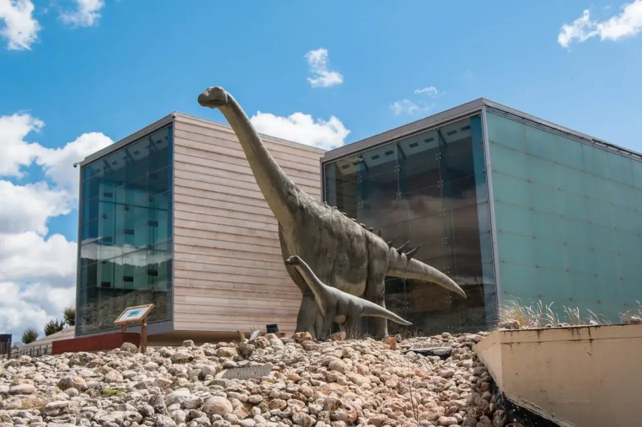 Dos dinosaurios, uno grande y uno pequeño, posan frente a edificios modernos. Un lugar ideal para una cuenca con niños.