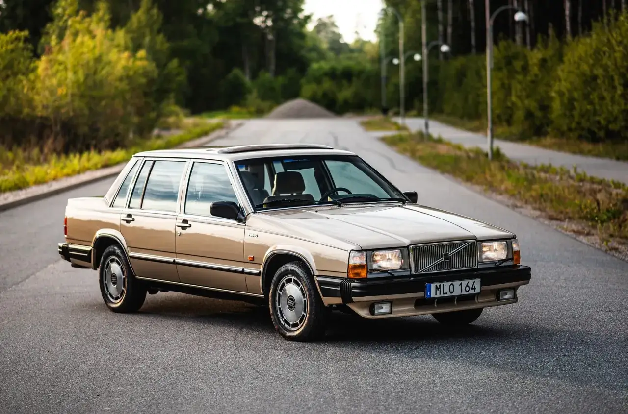En beige 760 Volvo st&aring;r parkerad p&aring; en v&auml;g kantad av tr&auml;d.