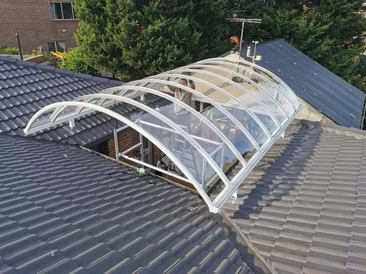 A curved, transparent canopy with a white frame is installed on a tiled roof. The canopy's flexible HDPE material allows for its arched shape, providing shelter.