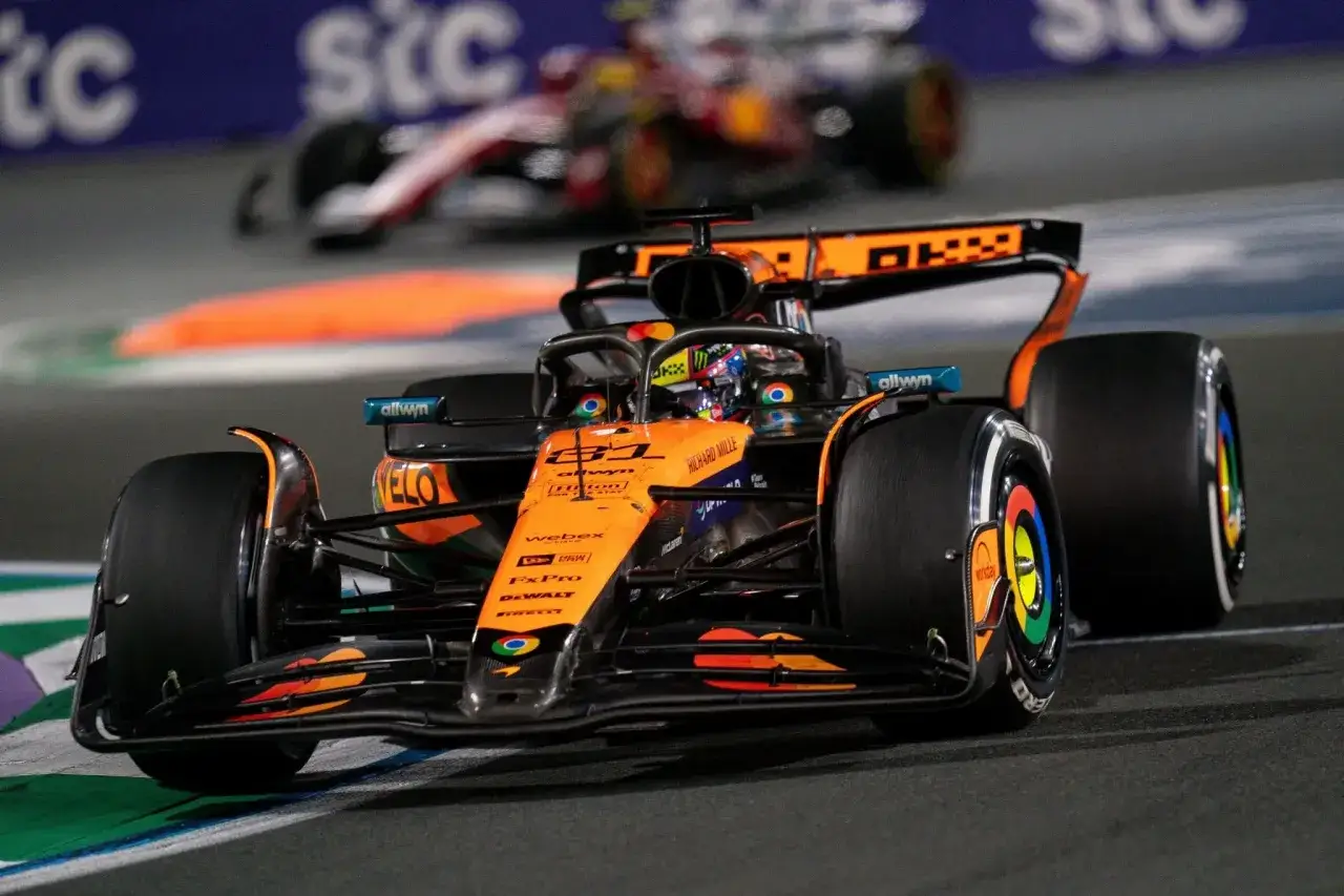 Pomarańczowy bolid F1 z numerem 81 na torze, walczący o cenne f1 punkty. W tle inny samochód.