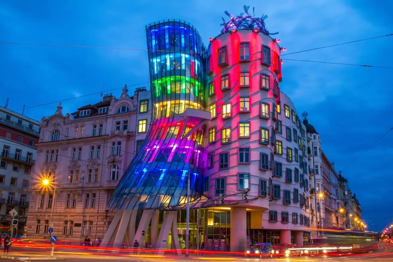 La maison dansante de Prague, illuminée de couleurs vives, semble danser sous le ciel nocturne. Les lumières des voitures créent des traînées lumineuses sur la rue.