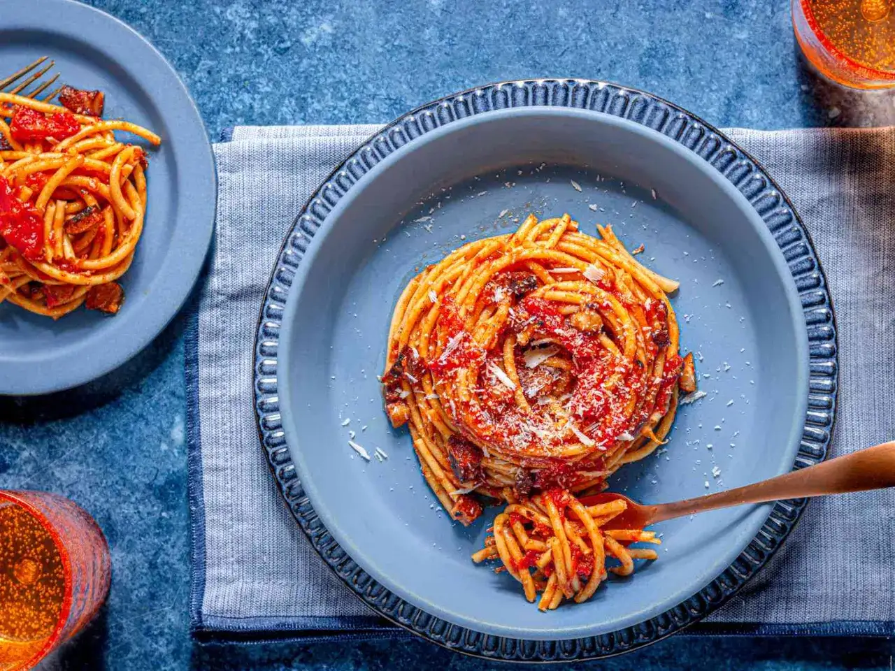 En portion bucatini all'amatriciana serveras i en blå skål, toppad med riven ost. En gaffel med pasta lyfts ur skålen.