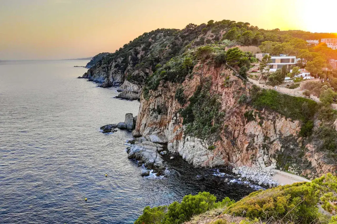 Un camino de ronda serpentea por acantilados rocosos y vegetación exuberante, con el mar Mediterráneo al atardecer.
