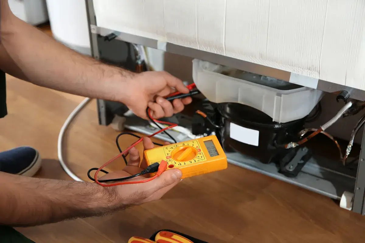 Un technicien vérifie le compresseur d'un frigo qui claque avec un multimètre.