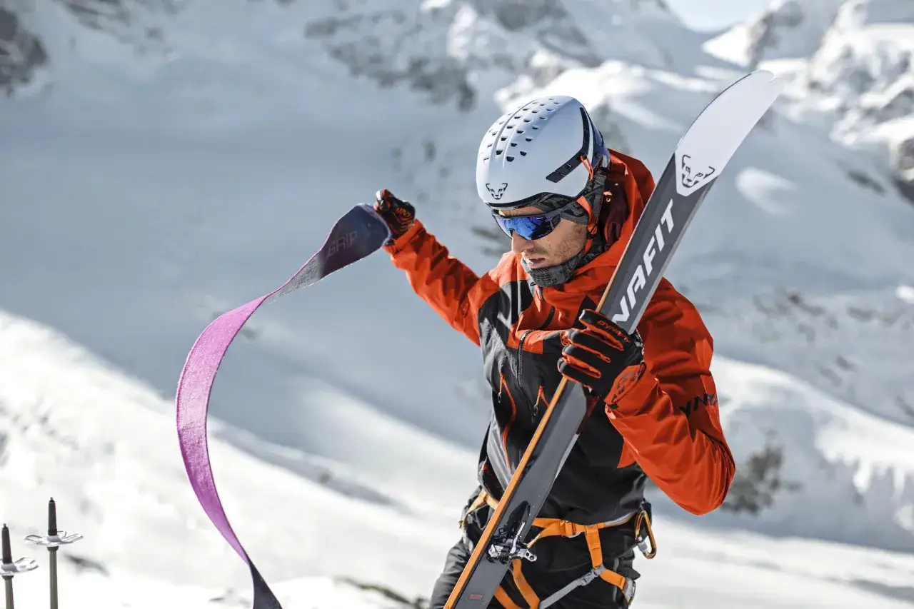 Mężczyzna w pomarańczowej kurtce przygotowuje się do skitury, trzymając narty i foki. To właśnie skitury co to za sport?