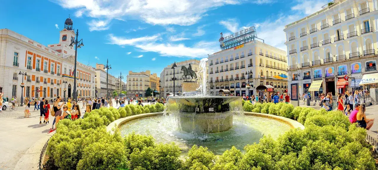 Tętniący życiem plac Puerta del Sol w Madrycie z fontanną i ludźmi spacerującymi pod błękitnym niebem.