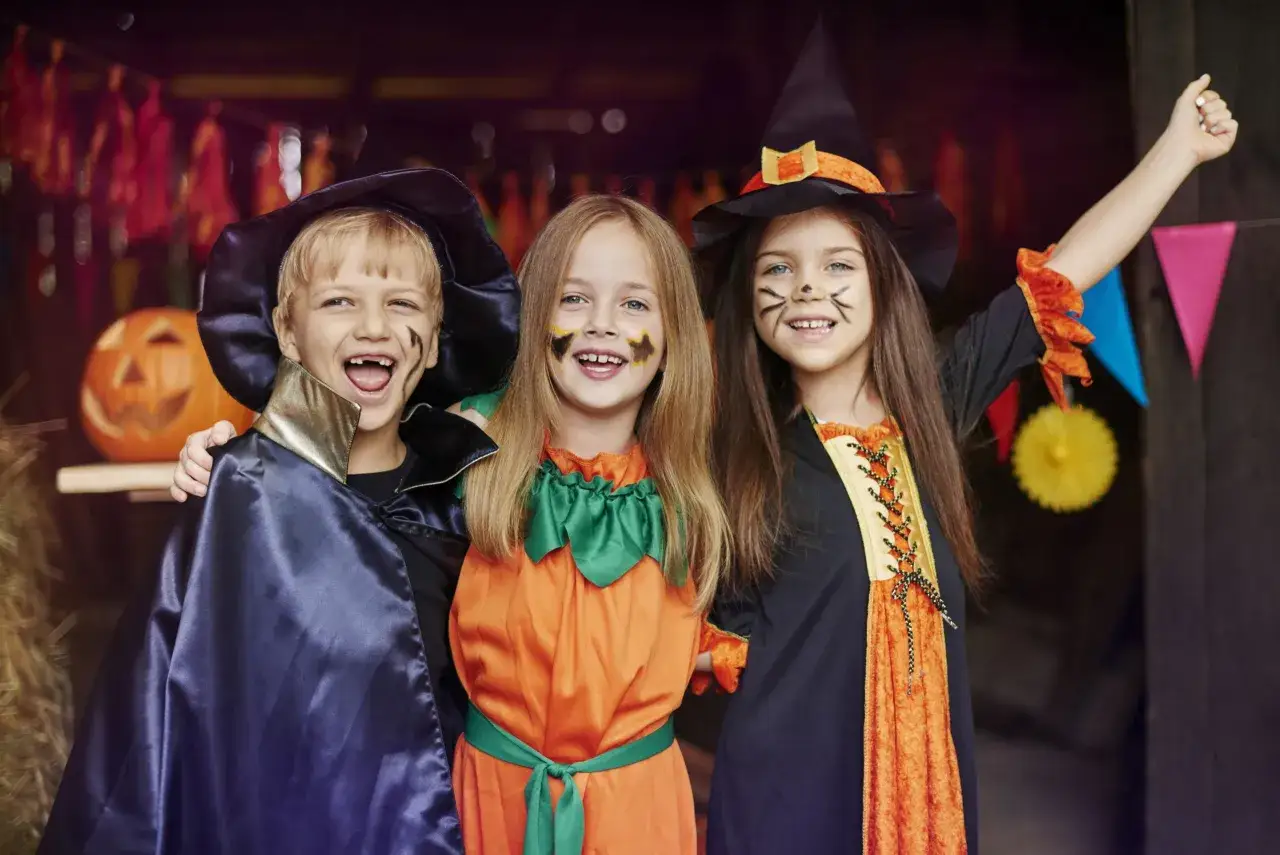 Tr&oacute;jka dzieci w strojach na Halloween: chłopiec w pelerynie, dziewczynka w stroju dyni i dziewczynka czarownica. Wesołe zagadki na Halloween!