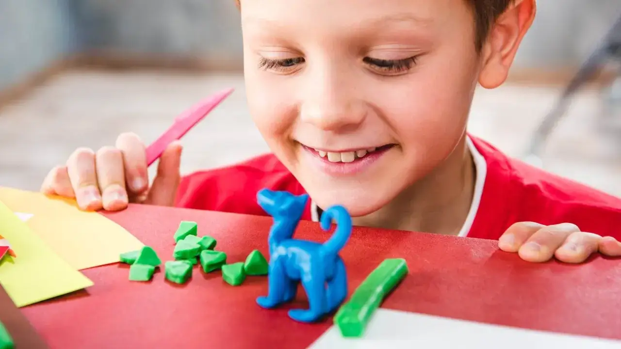 Ni&ntilde;o sonriente crea un dinosaurio azul con plastilina casera, rodeado de piezas verdes y herramientas de manualidades.