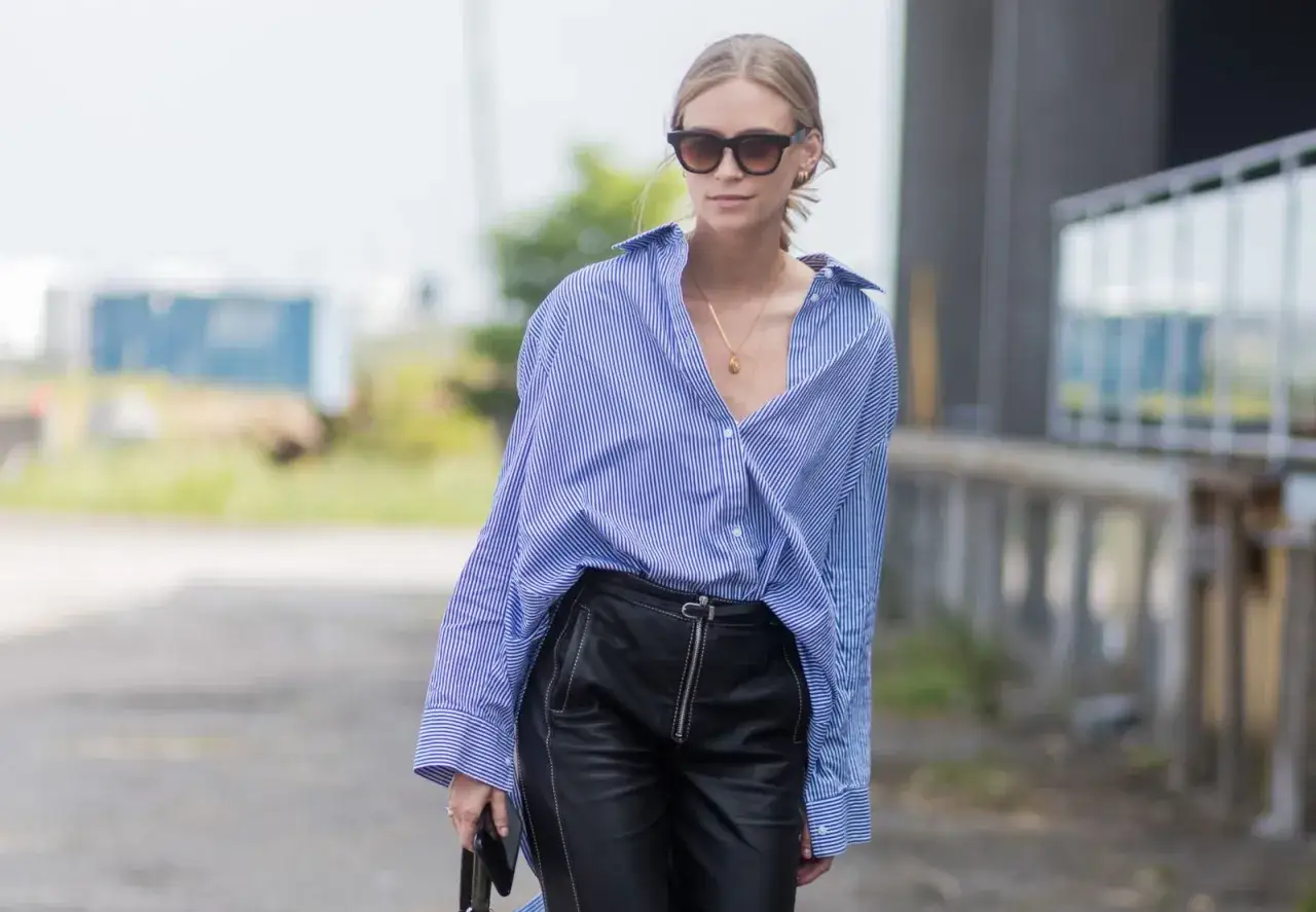 Mujer con camisa a rayas azules y blancas, pantalones de cuero negro y gafas de sol, luciendo un elegante french tuck.