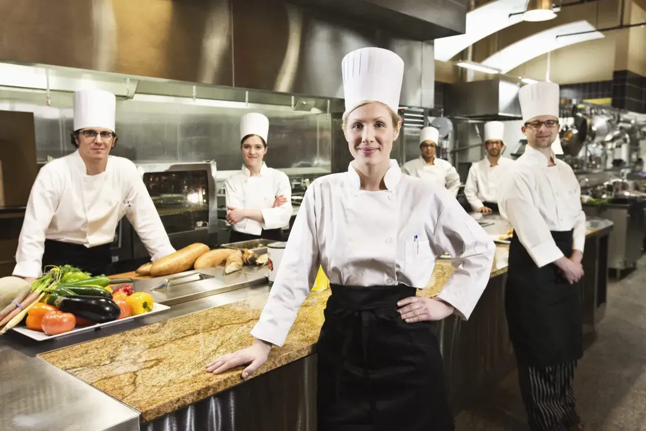 Chefs en formación en cursos de cocina para profesionales, sonriendo en una cocina profesional con verduras frescas y pan en la encimera.