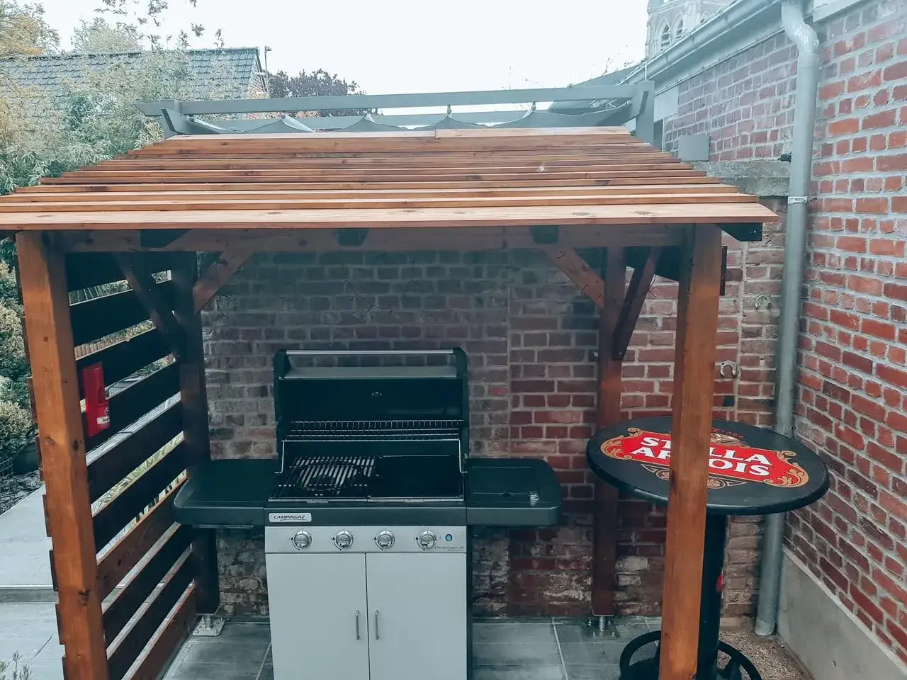 Un coin barbecue couvert, idéal pour fabriquer un espace convivial. Un barbecue, une table haute Stella Artois et une structure en bois.