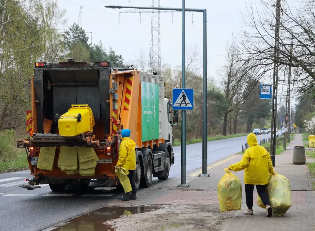 Pracownicy w żółtych płaszczach odbierają worki ze śmieciami do śmieciarki. To część umowy na wywóz śmieci.