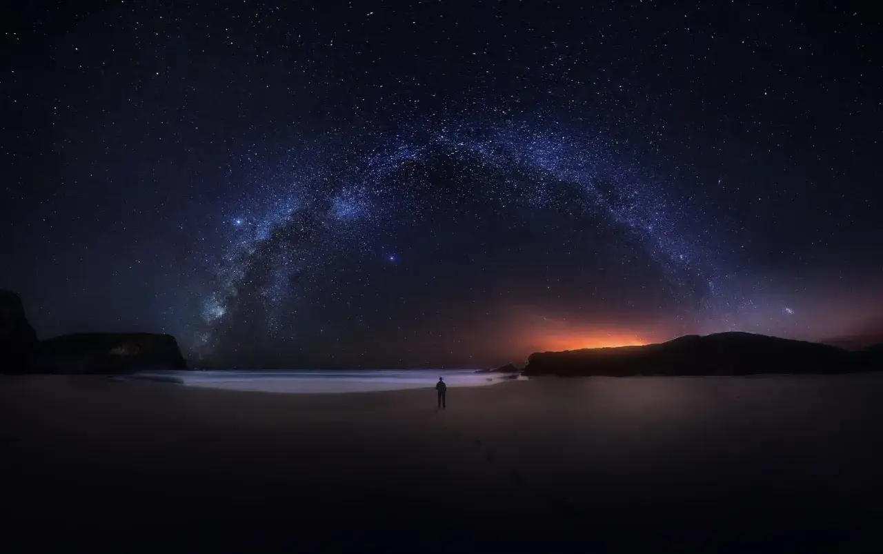 Samotna postać na plaży pod rozgwieżdżonym niebem, gdzie Droga Mleczna tworzy łuk. W oddali pomarańczowa łuna. **Pluton w strzelcu** inspiruje do nocnych wędrówek.
