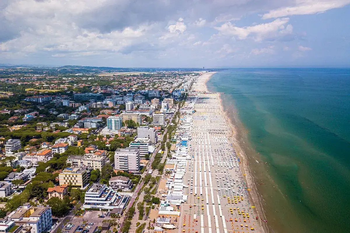 Vista aerea di Cattolica: una lunga spiaggia di sabbia con ombrelloni bianchi e gialli si estende lungo il mare Adriatico. La città si sviluppa alle spalle della costa.