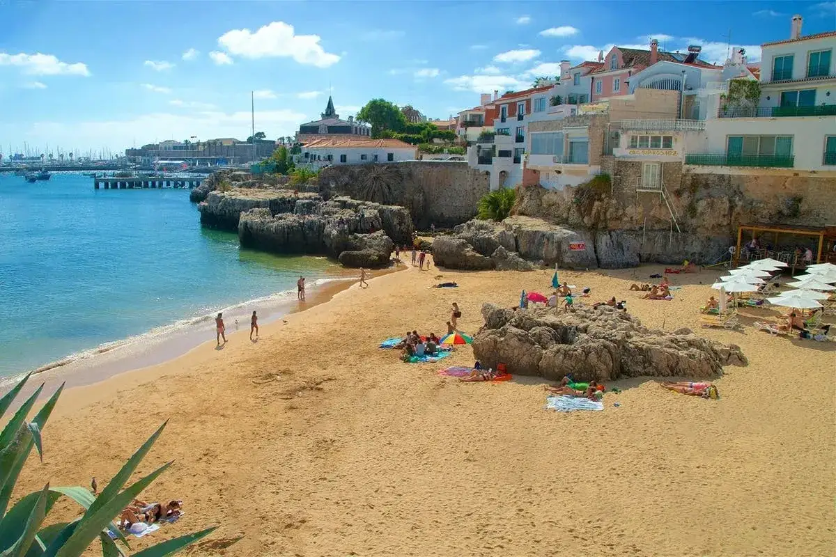 Sonniger lissabon strand mit Menschen, die sich am Sand und im Meer entspannen, im Hintergrund wei&szlig;e H&auml;user auf Felsen.