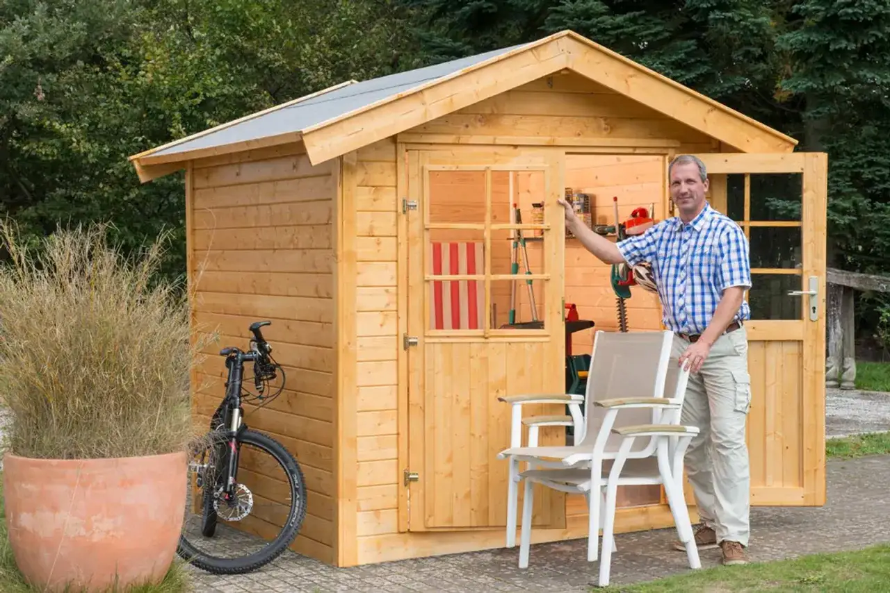 Mann steht vor einem neuen Gartenhaus, bereit, es zu streichen. Werkzeuge und ein Fahrrad sind bereits verstaut.