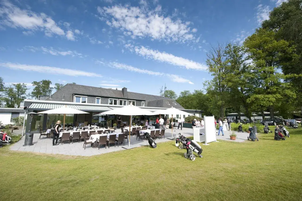 G&auml;ste genie&szlig;en das Bergisch Land Golf Resort bei Sonnenschein. Tische sind auf der Terrasse gedeckt, Golftaschen stehen bereit.