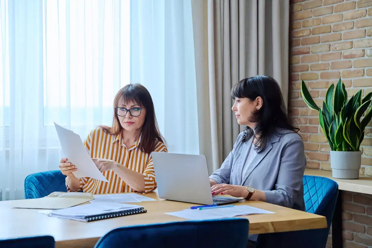 Two women in an office setting, one reviewing documents, the other on a laptop. This scene embodies effective presence coaching, fostering collaboration and growth.